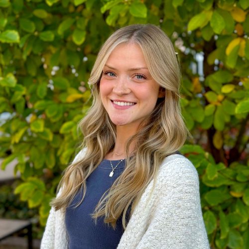 Young blonde woman wearing a white sweater smiles in front of a leafy green background.