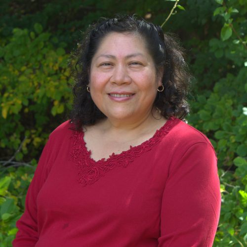 A woman with a red shirt and dark hair smiles in front of a large green bush.