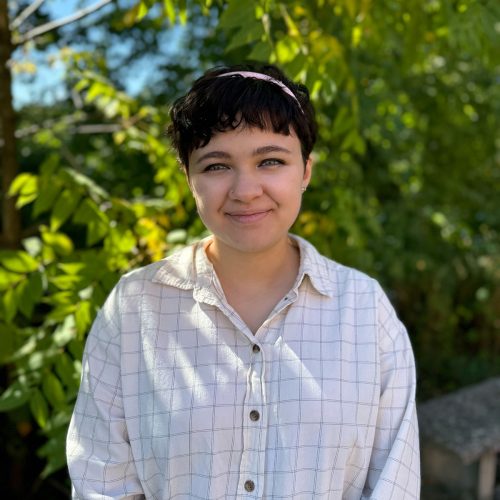 A young woman with short dark hair and a checkered shirt smiles in front of a tree lined park setting.