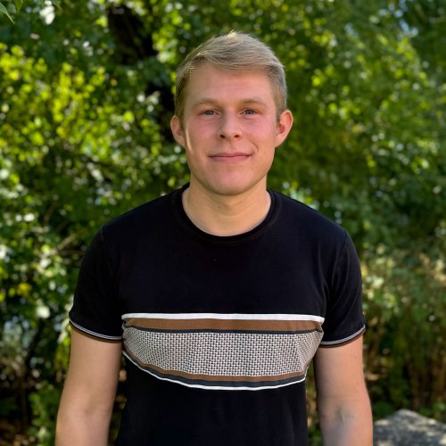 A blonde young man with a striped shirt smiles in front of a leafy background.