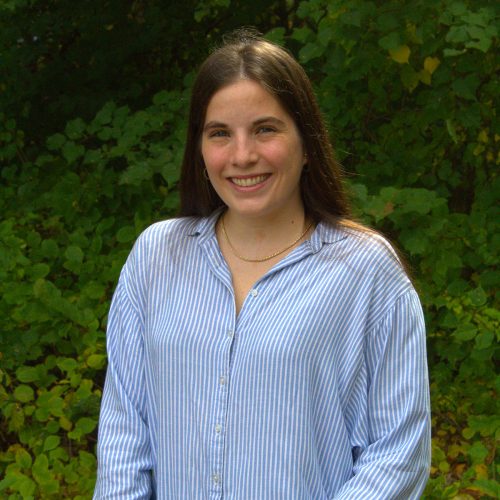 A woman with long dark hair and a blue stripe shirt smiles in front of lush dark green trees.