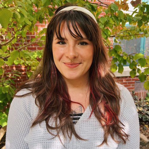 A young woman with long brown hair and red highlights, smiles against a leafy tree and brick background.