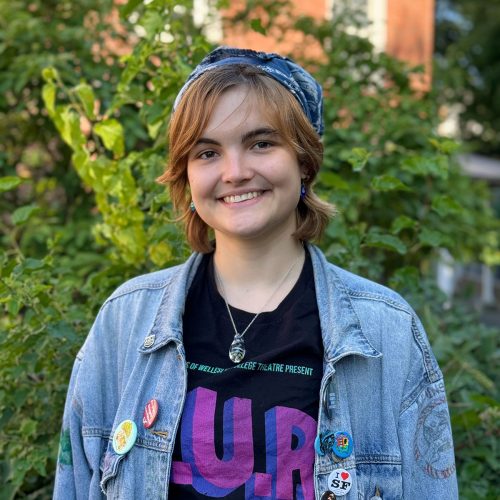 A young woman with a kerchief on her head, and black t-shirt smiles in front of a green forest of trees.