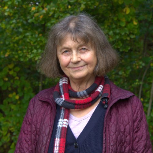 An elderly woman wears a scarf and purple coat in front of a green forest.