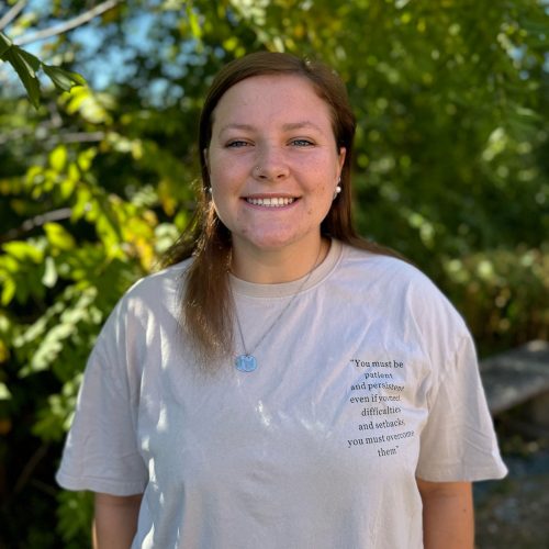 A young woman in a gray t-shirt smiles in front of lush, green trees.