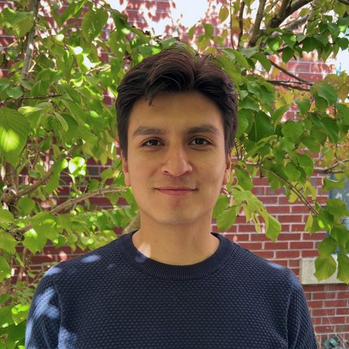 A man with brown hair and olive skin smiles in front of a leafy green tree in the background.